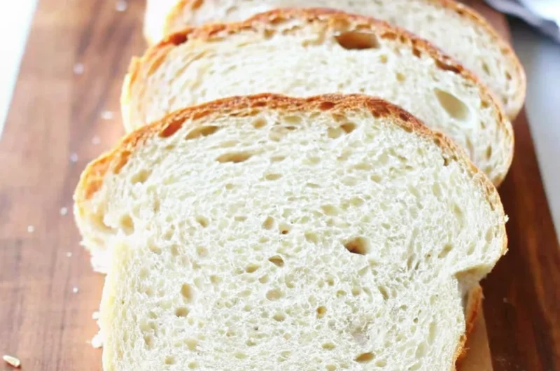 A freshly baked loaf of homemade sandwich bread with a golden-brown crust, resting on a wooden cutting board with a few slices cut to reveal its soft texture.