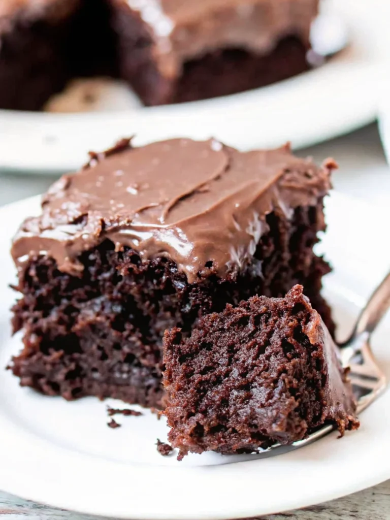 Moist chocolate zucchini brownie topped with rich chocolate frosting on a white plate, with a fresh zucchini in the background.