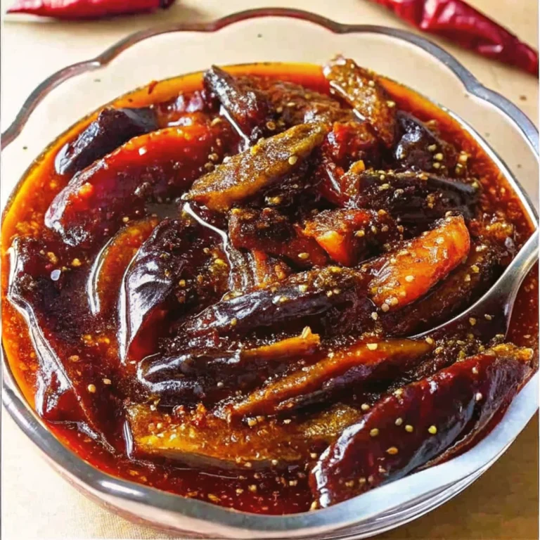 A bowl of spicy Indian brinjal pickle (eggplant achar) in oil with visible mustard seeds and red chili flakes.
