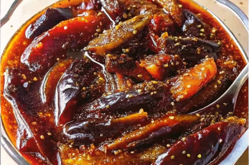 A bowl of spicy Indian brinjal pickle (eggplant achar) in oil with visible mustard seeds and red chili flakes.