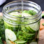 A close-up of a glass jar filled with freshly made refrigerator pickles, featuring sliced cucumbers, fresh dill sprigs, garlic cloves, and spices in a tangy brine—perfect for an easy dill pickles recipe.