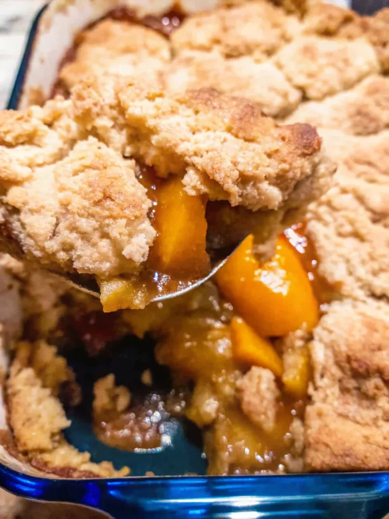 A bowl of Southern peach cobbler with fresh peaches, topped with golden biscuit crust, served on a rustic wooden table with a vintage spoon.