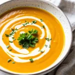 A bowl of creamy pumpkin soup garnished with fresh parsley, a swirl of cream, and cracked black pepper.