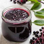 Homemade elderberry syrup recipe in glass jar with fresh elderberries, cinnamon stick, and honey on wooden background