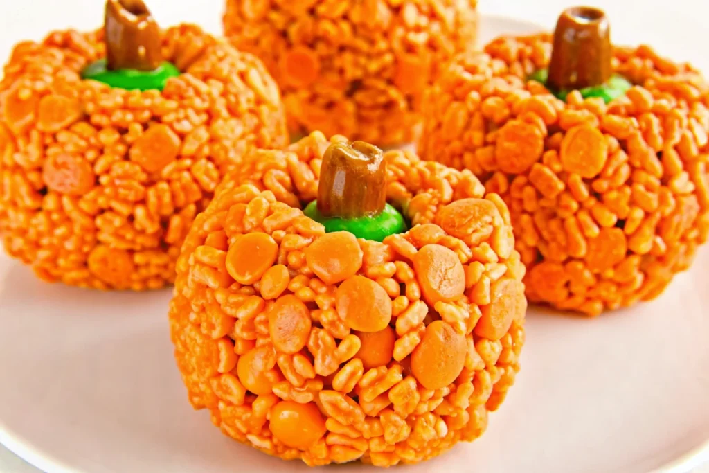 Orange pumpkin-shaped Rice Krispie treats with brown Tootsie Roll stems and green M&M leaves arranged on white background for Halloween party dessert