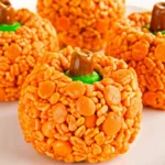 Orange pumpkin-shaped Rice Krispie treats with brown Tootsie Roll stems and green M&M leaves arranged on white background for Halloween party dessert
