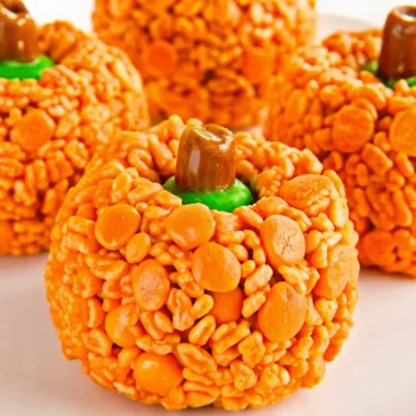Orange pumpkin-shaped Rice Krispie treats with brown Tootsie Roll stems and green M&M leaves arranged on white background for Halloween party dessert