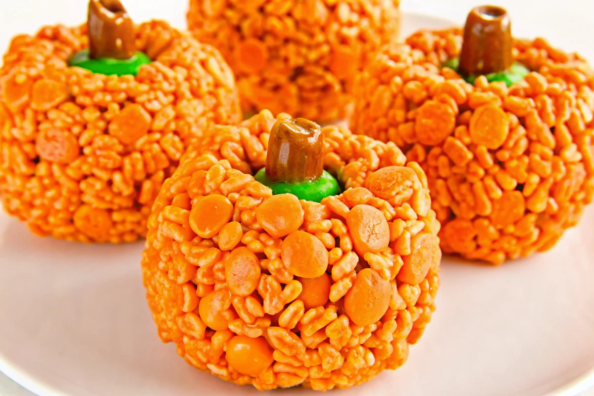 Orange pumpkin-shaped Rice Krispie treats with brown Tootsie Roll stems and green M&M leaves arranged on white background for Halloween party dessert