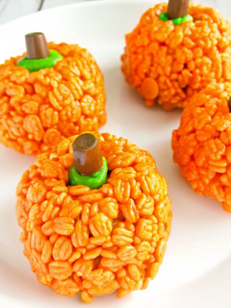 Orange pumpkin-shaped Rice Krispie treats with brown Tootsie Roll stems and green M&M leaves arranged on white background for Halloween party dessert