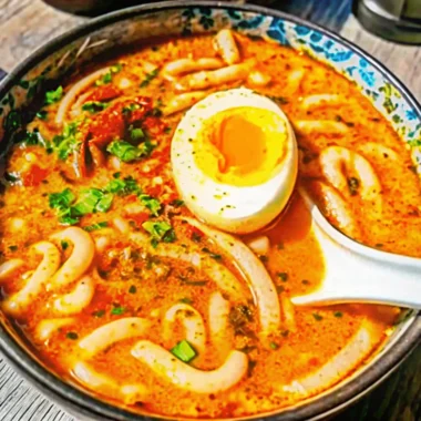 Steaming bowl of spicy peanut udon noodle soup with thick white udon noodles in creamy peanut broth, topped with soft-boiled egg, green onions, and sesame seeds