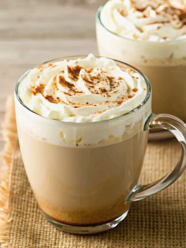 Homemade Pumpkin Spice Latte in clear glass mug with whipped cream topping and cinnamon stick garnish on wooden table with fall leaves