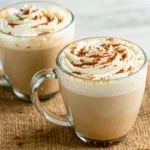 Homemade Pumpkin Spice Latte in clear glass mug with whipped cream topping and cinnamon stick garnish on wooden table with fall leaves