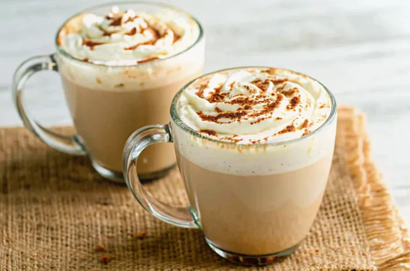 Homemade Pumpkin Spice Latte in clear glass mug with whipped cream topping and cinnamon stick garnish on wooden table with fall leaves